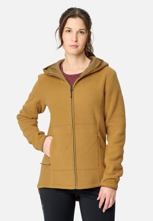 Junge Frau mit braunen Haaren, die eine senffarbene Fleecejacke mit Reißverschluss und Kapuze sowie schwarze Hose trägt und mit einer Hand in der Tasche posiert.