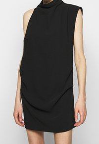 Svart ärmlös klänning med draperad design, hög krage och mjukt tyg. Slät textur och knälång cut. Inga synliga dekorationer.