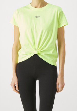 Femme portant un T-shirt manches courtes jaune fluo noué à la taille avec le logo "DKNY" et un pantalon noir ajusté, sur un fond uni.