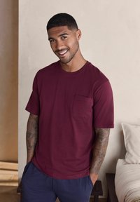 Uśmiechnięty mężczyzna o krótkich włosach w bordowej koszulce i granatowych spodniach, stojący z rękami w kieszeniach obok łóżka i jasnej ściany.