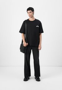 Black oversized t-shirt with short sleeves, detailed with a white graphic on the left chest, paired with dark wide-leg jeans and black shoes.