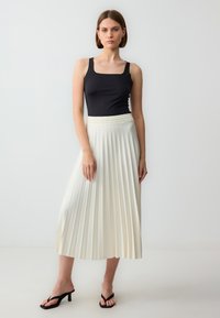 Zwarte mouwloze top en een lange witte geplooide rok met een elastische tailleband, gecombineerd met zwarte hoge sandalen. Glad stof en strakke lijnen.