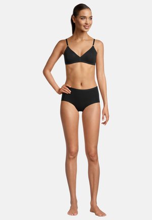 Frau steht barfuß, trägt einen schwarzen BH und hoch taillierte schwarze Unterwäsche, lächelt und schaut zur Seite, mit einer Hand auf der Hüfte.