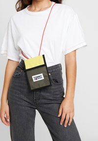 Borsa a tracolla verde e gialla con una tracolla rossa, con bordi neri e logo "TOMMY JEANS". Il materiale sembra resistente con una texture liscia.