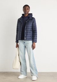 Giacca di piumino blu navy con cuciture orizzontali e bottoni a pressione, abbinata a una felpa scura e jeans a gamba larga di un blu chiaro. Tiene una borsa a mano color crema.