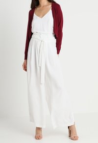 White wide-leg trousers with a tied belt, paired with a burgundy cardigan and a white camisole. The outfit is completed with nude heeled sandals.