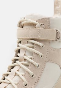 Primo piano di una sneaker alta beige che mostra la texture del tessuto, i lacci rotondi, gli occhielli, i pannelli in suede cuciti e la tracolla regolabile con velcro.