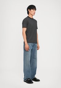 T-shirt noir à motifs avec un col côtelé, associé à un jean en denim bleu ample et des chaussures noires, présenté sur un fond neutre.