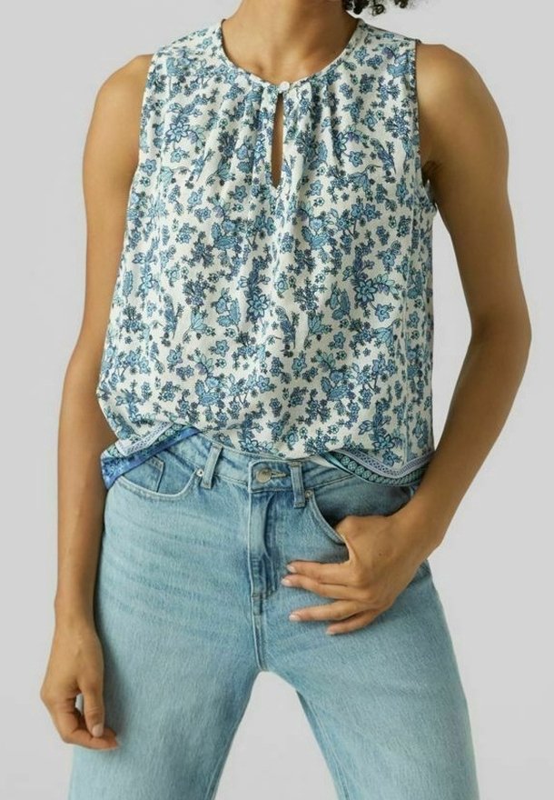 Femme portant un haut blanc sans manches avec un imprimé floral bleu, rentré dans un jean bleu clair taille haute, une main dans la poche et l'autre sur le côté.