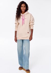 Beige mikina s růžovými šňůrkami, velká přední kapsa a detail loga. V kombinaci s světle modrými širokými džíny a hnědými botami.