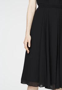 Schwarzes Kleid mit einem gefältelten Design, Taillendetails und transparenter Textur. Der Saum reicht bis unter das Knie. Eine Hand ist teilweise sichtbar.