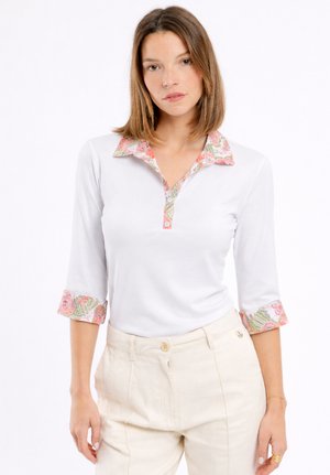 Femme portant une chemise blanche avec un bord à fleurs sur le col et les manches, associée à un pantalon crème taille haute, debout devant un fond blanc.