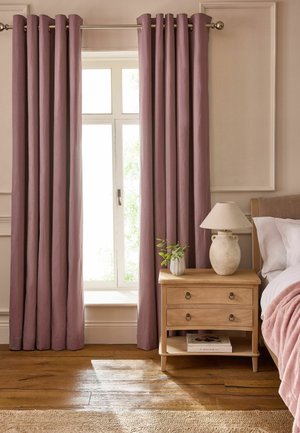 Finestra con tende malva accanto a un comodino in legno con una lampada beige, una piccola pianta e un libro, accanto a un letto con cuscini bianchi e una coperta rosa.