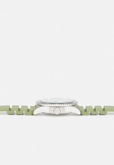 Montre-bracelet avec un boîtier en acier inoxydable argenté, un verre transparent et un bracelet en silicone vert en zigzag. Cadran rond avec des marqueurs d'heure numériques.