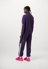 Violet sportinis treningas su baltais akcentų dryžiais, turintis užtrauktuką striukę ir atitinkančias kelnes. Ryškiai rožiniai sportiniai bateliai užbaigia aprangą.