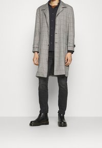 Grå rutig kappa med krage, knäppning fram och justerbara ärmmuddar, bärs över en mörk skjorta och slim fit svarta byxor med svarta stövlar.