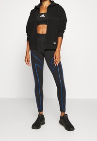 Sudadera negra con cremallera y hombros texturizados, sujetador deportivo negro, leggings negros con rayas azules y zapatos deportivos negros.