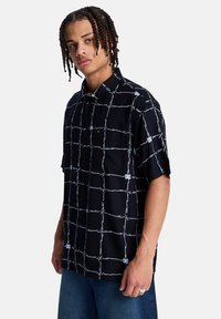 Μαύρο πουκάμισο με κοντά μανίκια και κουμπιά, με λευκό σχέδιο πλέγματος και διακοσμητικά σύρματα με αγκάθια, διαθέτει χαλαρή εφαρμογή και λεία υφή υφάσματος.