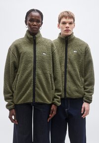 To personer bærer olivengrønne fleecetrøjer med høje kraver, lynlåse foran og sidelommer. Trøjerne har et lille logo på brystet.