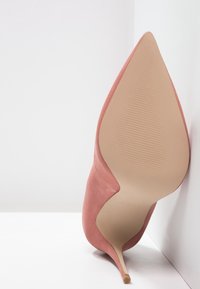 Chaussure à talon haut en suede rose avec un bout pointu, des côtés découpés et un talon aiguille fin. Texture lisse et détails de design minimalistes.