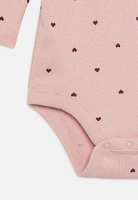 Pelele de algodón rosa claro para bebés con pequeños patrones de corazones color burdeos. Presenta mangas largas y cierre a presión en la parte inferior para facilitar el vestido.