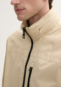 Beige lichte jas met hoge kraag, zwarte rits en gestikte details. Bevat een zijzak met een subtiel decoratief element.