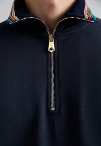 Sweat-shirt zippé bleu marine avec col rayé coloré et fermeture éclair dorée équipée d'un tire-zip en forme de triangle. Le matériau est lisse avec une finition texturée.