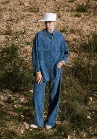 Blauwe textuurdenimshirt met twee borstzakken, gecombineerd met bijpassende wijde broek. Heeft een brede witte hoed en gouden accessoires.