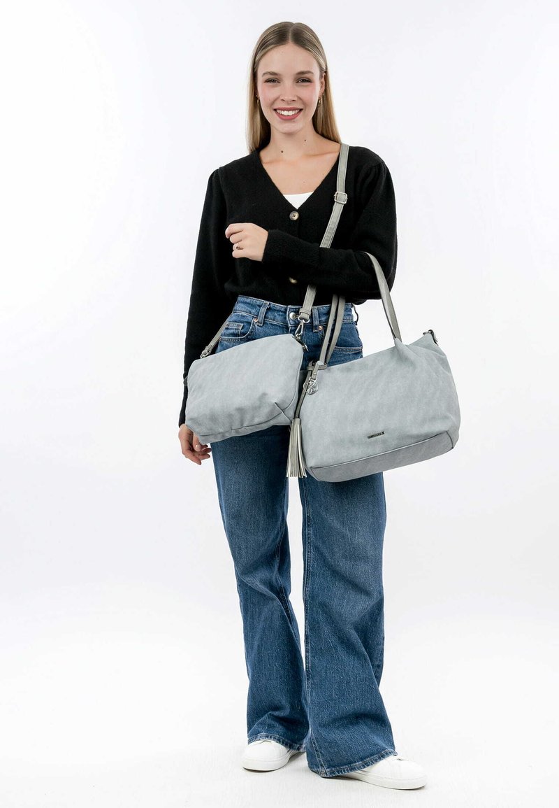 Gray handbag with a soft texture and a tassel detail, featuring a roomy design and a shoulder strap, paired with a black cardigan and jeans.