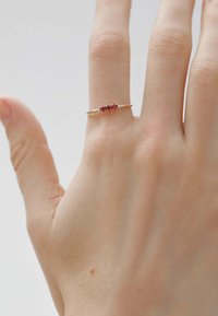 Anillo de oro con cinco pequeñas piedras preciosas rojas, adornado con dos acentos claros, descansando sobre una mano de tono de piel claro con un fondo neutral.