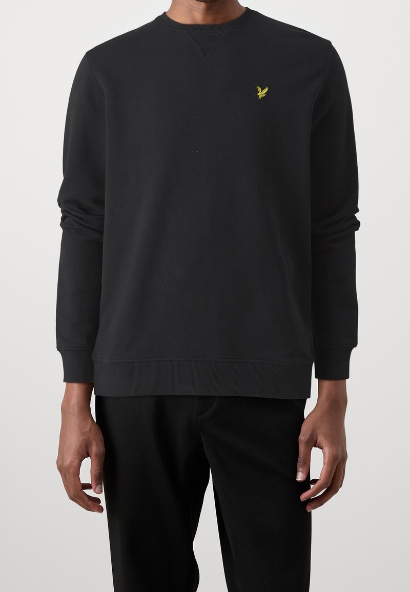 Zwarte crewneck sweater van zacht fabric, met een klein geel embleem op de linker borst en geribde boorden en onderrand.