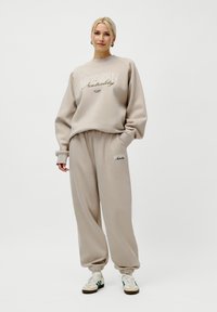 Kvinne står iført beige genser og matchende joggebukser med teksten "Modern Neutrality", kombinert med hvite joggesko.