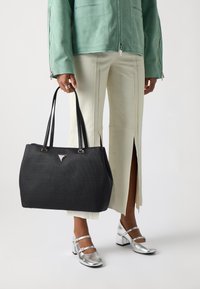 Geantă de umăr din material textil negru, cu formă structurată, mânere din piele, accesorii din argint și un accent logo triangular. Asortată cu pantaloni deschiși la culoare și pantofi argintii.