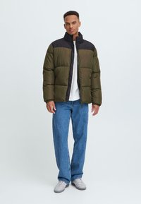 Giacca a vento in verde oliva con una parte superiore nera, caratterizzata da un colletto alto, zip frontale e texture trapuntata visibile. Indossata con jeans blu.