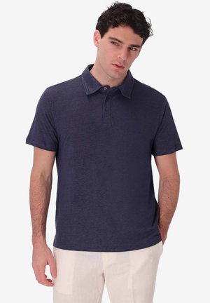 Uomo con capelli scuri e ricci che indossa una polo blu scuro a maniche corte e pantaloni beige chiaro, in piedi davanti a uno sfondo semplice.
