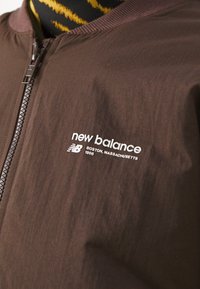 Hnědá zipová bunda s bílým logem "new balance Boston, Massachusetts 1906" na hrudi, oblečená přes černé a žluté pruhované tričko.