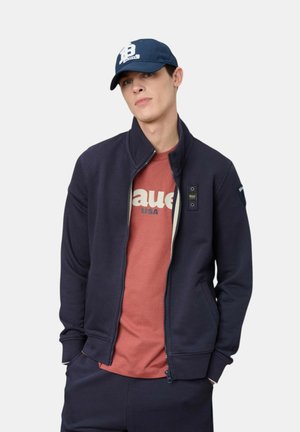 Giovane uomo con giacca blu scuro con cerniera sopra una maglietta rossa con logo e un cappellino da baseball blu scuro, in piedi con le mani in tasca.
