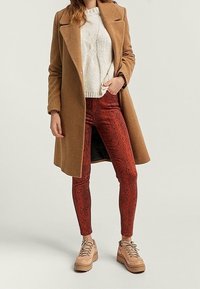 Bruin wollige overjas, crèmekleurige gebreide trui, rode slangenprint skinny broek en beige platform sneakers met veters.