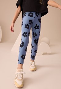 Kék leggings fekete virágmintával, testhezálló kialakítású és rugalmas anyagból. Fehér és sárga tornacipővel párosítva.