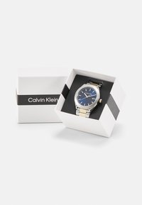 Reloj Calvin Klein en una caja blanca, con una pulsera de acero inoxidable en tono plateado y acentos en dorado, un dial azul y una pantalla de fecha.