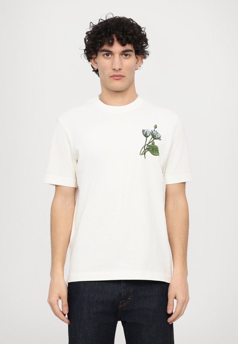 Cremefarbene Baumwoll-T-Shirt mit kurzen Ärmeln und Rundhalsausschnitt, ausgestattet mit blauer Blumenstickerei auf der linken Brust.