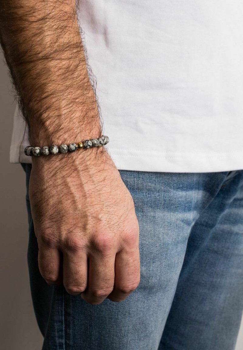 Pelēka un melna raibā akmens pērlīšu aproce ar zelta akcentu, uzlikta uz rokas, valkāta kopā ar baltu T-kreklu un zilām džinsu biksēm.