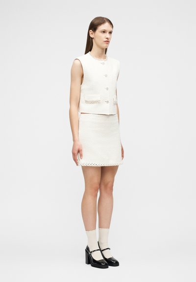 Jeune femme debout de profil portant un gilet sans manches blanc texturé, une jupe assortie, des chaussettes crème et des escarpins noirs style Mary Jane.