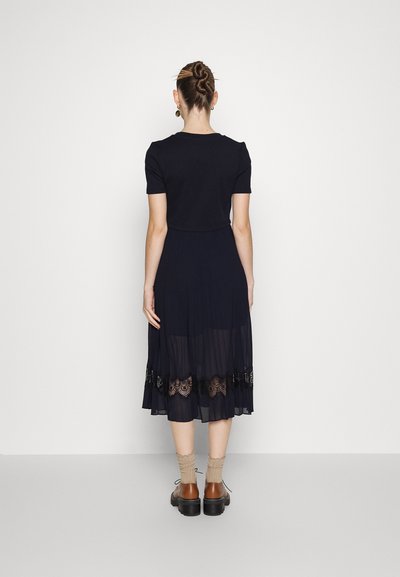 Vestido navy con mangas cortas y falda transparente, con un borde de encaje bordado en el dobladillo. Lleva zapatos marrones y calcetines claros.