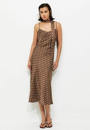 Femme debout contre un mur blanc, portant une robe midi marron sans manches à pois blancs avec une écharpe assortie et des sandales noires.
