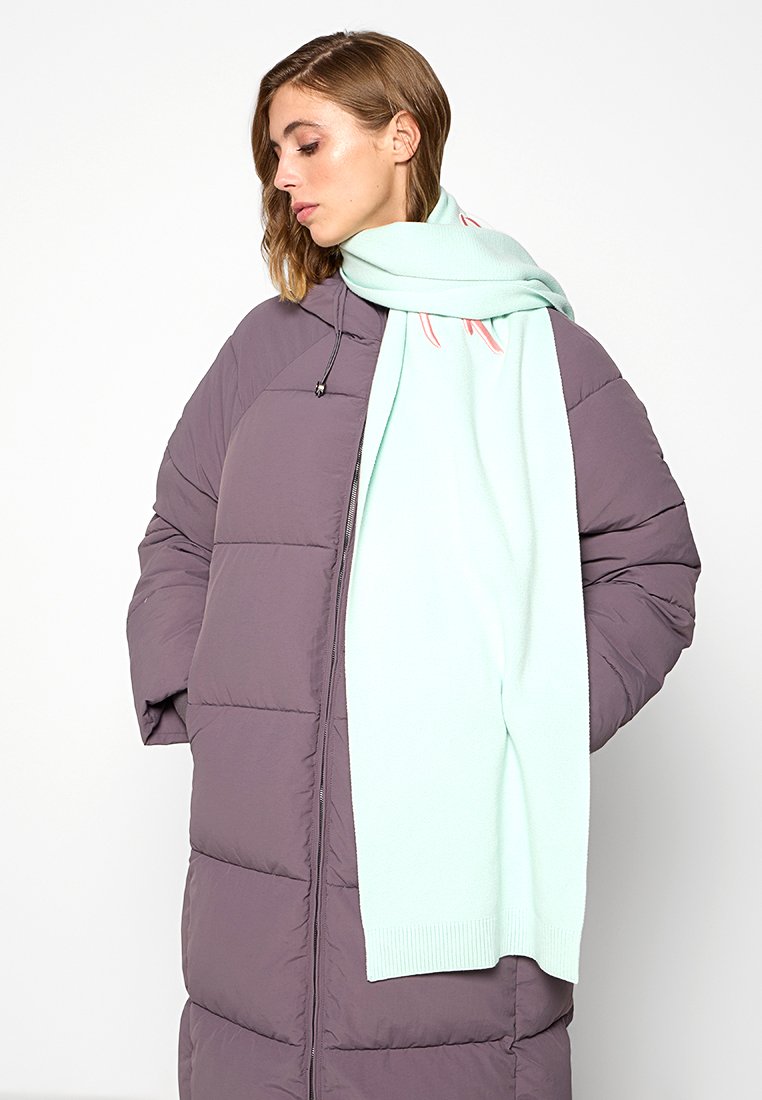 Ein hellblauer Schal liegt über einem lila gefütterten Daunenmantel mit Reißverschluss. Der Schal hat gerippte Enden und eine glatte Textur; der Mantel hat ein gestepptes Design.