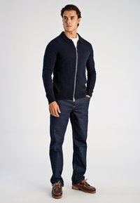 Granatowy sweter zapinany na zamek z szerokim kołnierzem, noszony na białej koszulce; zestawiony z ciemnymi dżinsami i brązowymi butami typu boat shoes. Gładka dzianina.