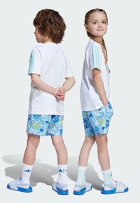 Vita t-shirts med gröna ränder på ärmarna, blå blomsterkortbyxor, vita strumpor och blåa halkskyddsandaler.