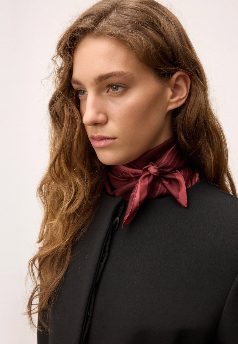 Palton negru cu guler înalt. Eșarfă de satin roșie legată la gât. Păr lung, ondulat. Machiaj natural cu tonuri subtile. Fundal neutru.