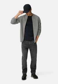 Grau gerippter Cardigan über einem schwarzen T-Shirt, kombiniert mit dunkelgrauen Jeans und schwarzen Schuhen. Das Modell trägt eine schwarze Mütze, die Hand zum Schirm erhoben.